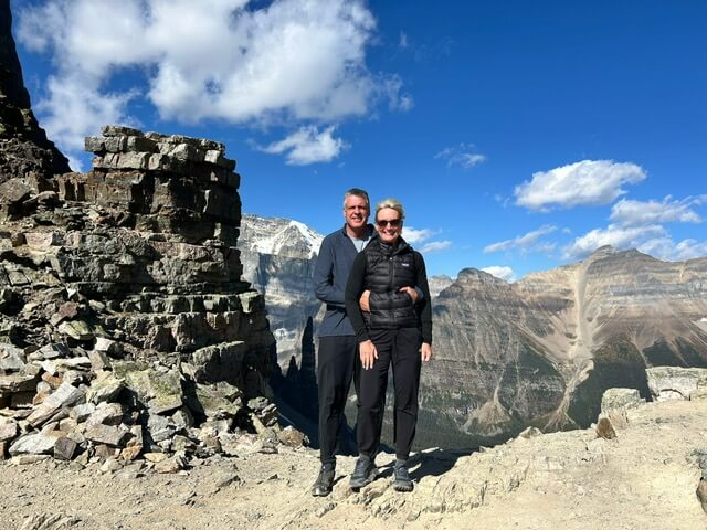 Paige and Bob on a high mountain trail with a stacked-rock cliff and snow-dusted peaks in Banff National Park